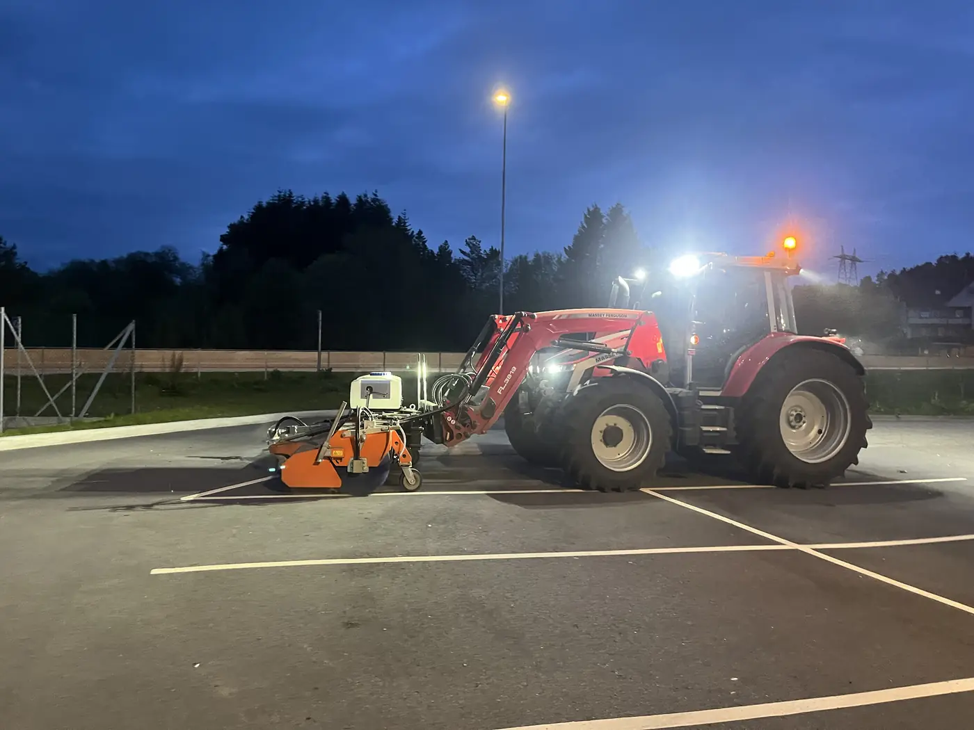 Traktor med redskap i arbeid på parkeringsplass om kvelden